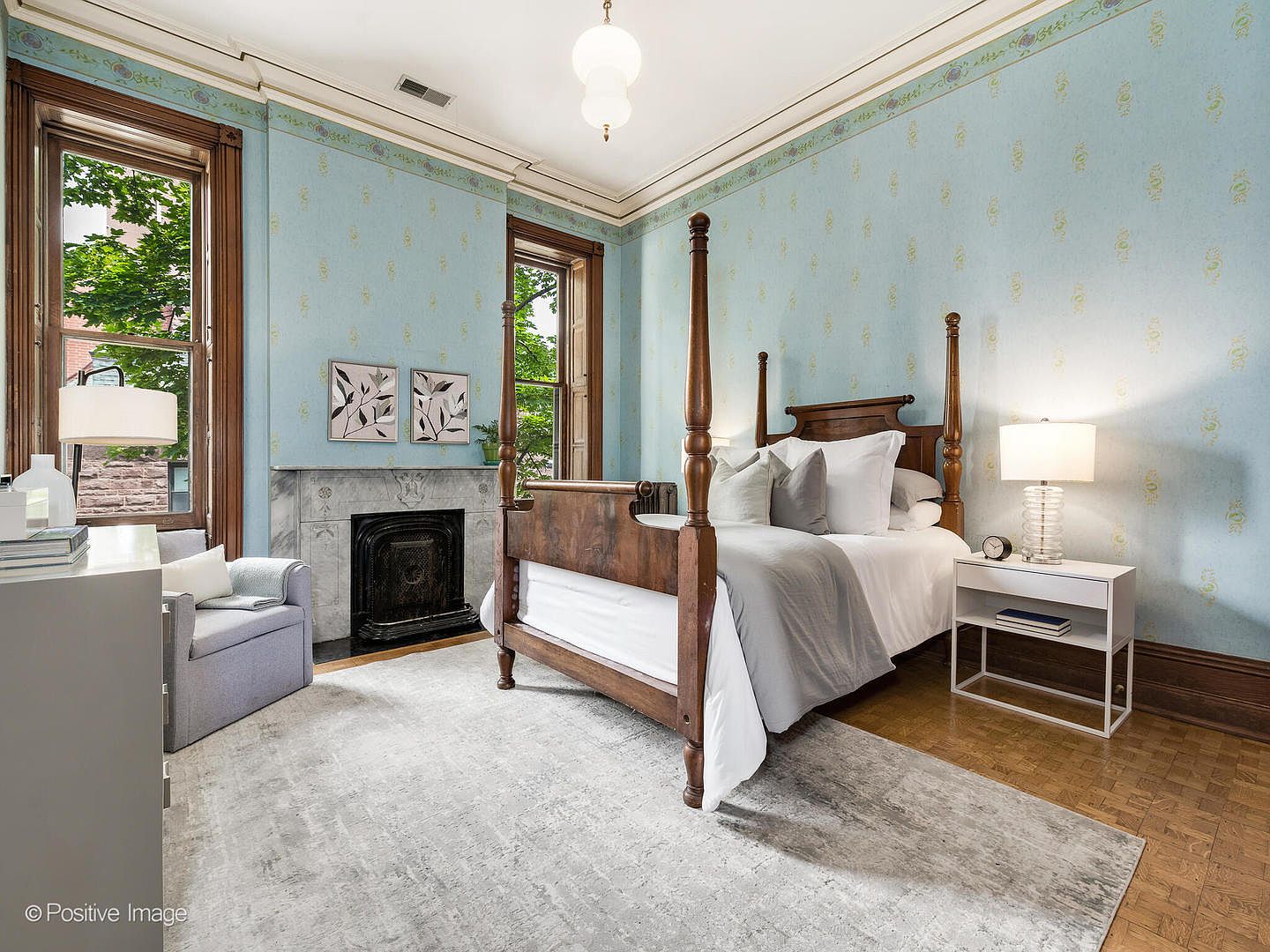 This elegant bedroom features light blue patterned wallpaper and classic wood trim, centered around a traditional four-poster bed. A stone fireplace with a decorative mantel serves as a focal point, flanked by two tall windows that allow for ample natural light. The room is finished with a large area rug over parquet wood flooring and a cozy armchair, creating a sophisticated and inviting atmosphere.