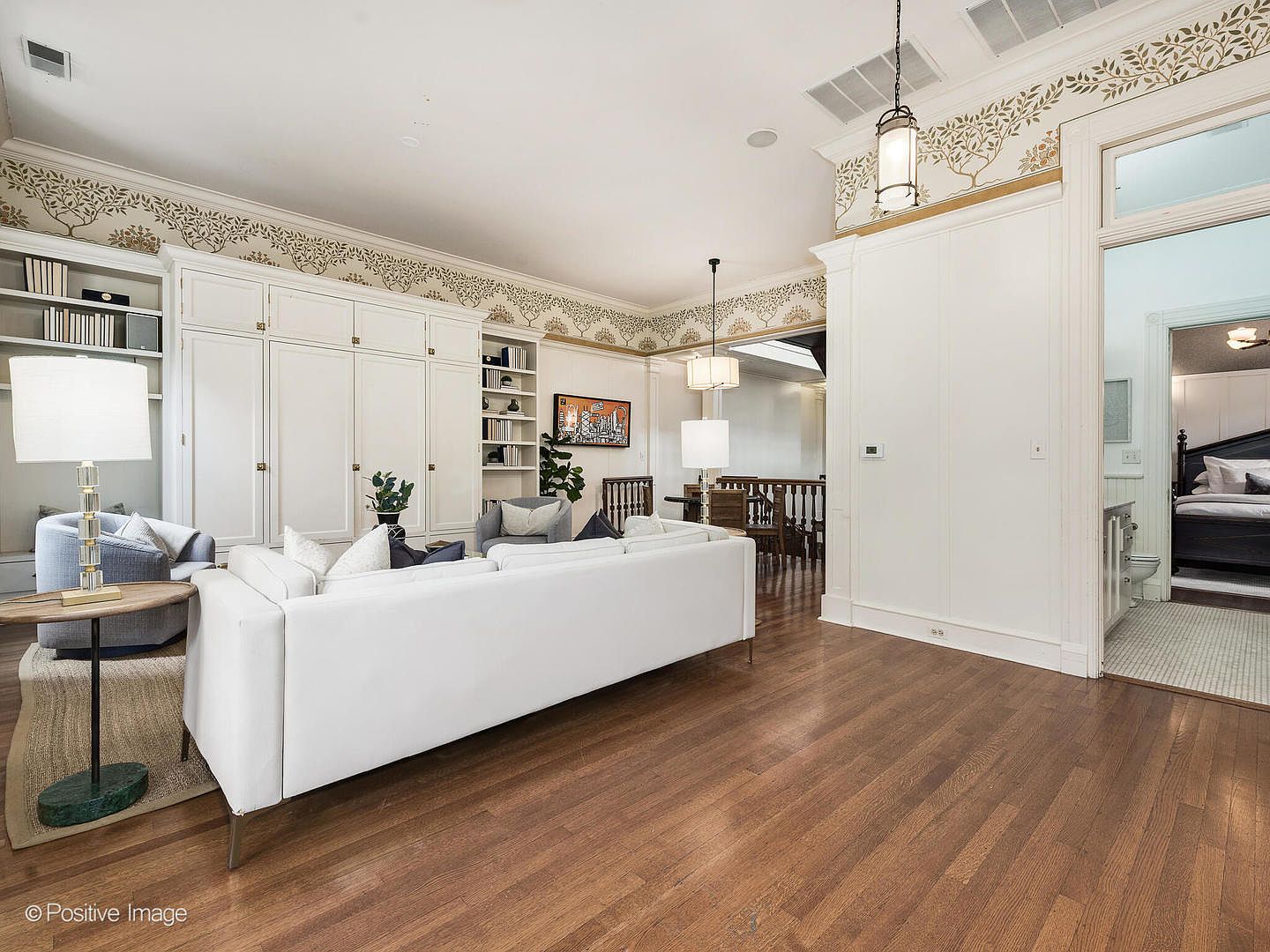 This bright and airy living room features a large white sofa as the centerpiece, complemented by a blue armchair and a round side table with a lamp. The room is characterized by extensive built-in white cabinetry and a decorative tree-patterned wallpaper border near the ceiling. A doorway on the right provides a glimpse into a bedroom, while the open layout flows toward a dining area in the background.