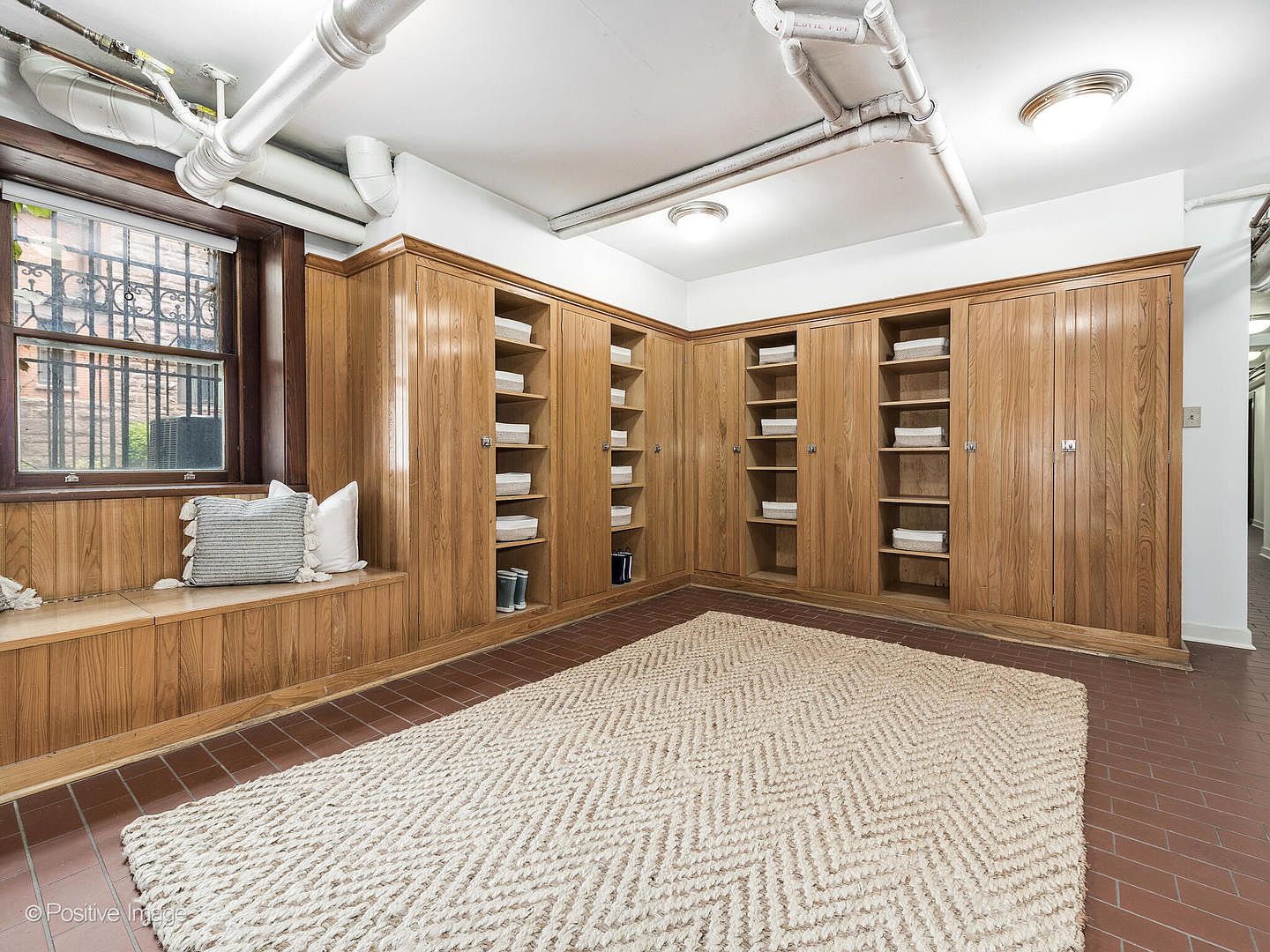 This basement space features extensive light wood built-in cabinetry and shelving units that wrap around the corner, providing ample storage. A built-in bench with plush pillows sits beneath a window with security bars, while a large, textured area rug covers the tiled floor. The ceiling is exposed with visible white piping, giving the room a functional, industrial-inspired aesthetic.