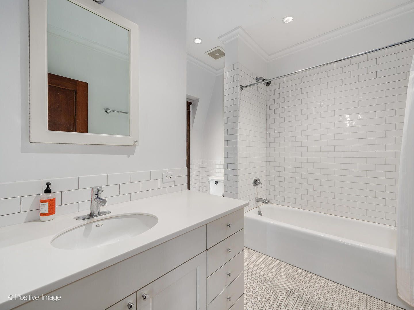 This bright, clean bathroom features a white vanity with a stone countertop and an oval undermount sink, paired with a rectangular mirror. A white subway-tiled shower-tub combination occupies the back of the room, complemented by classic white hexagonal floor tiles. The space is well-lit with recessed lighting and maintains a crisp, neutral aesthetic.