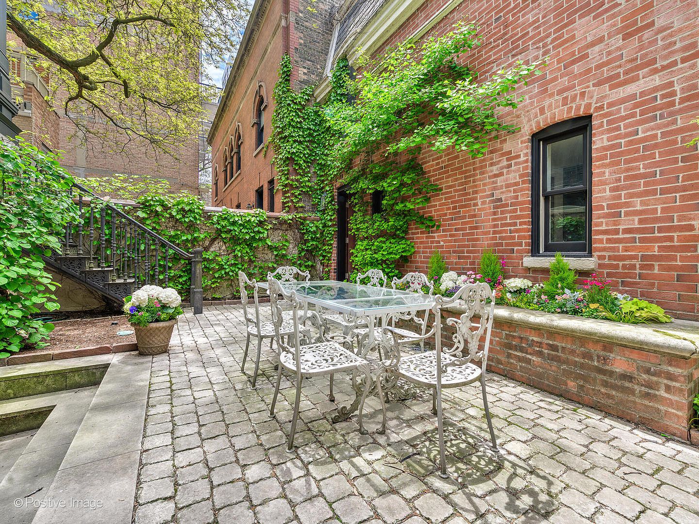 This charming outdoor patio features a classic wrought-iron dining set arranged on a cobblestone surface, set against a historic brick building adorned with climbing ivy. The space includes a raised brick planter filled with vibrant flowers and greenery, alongside an ornate metal staircase leading to an upper level. The perspective captures a serene, private courtyard atmosphere, perfect for al fresco dining in a sophisticated urban setting.