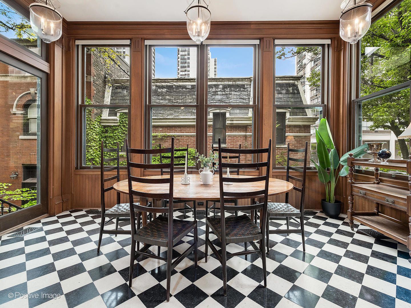 This elegant dining room features a classic black-and-white checkered floor and rich wood-paneled walls that create a sophisticated, traditional atmosphere. A round wooden table is centered in the room, surrounded by four high-backed chairs, with large windows providing ample natural light and views of the exterior greenery. Three glass pendant lights hang from the ceiling, adding a refined touch to this bright and inviting space.