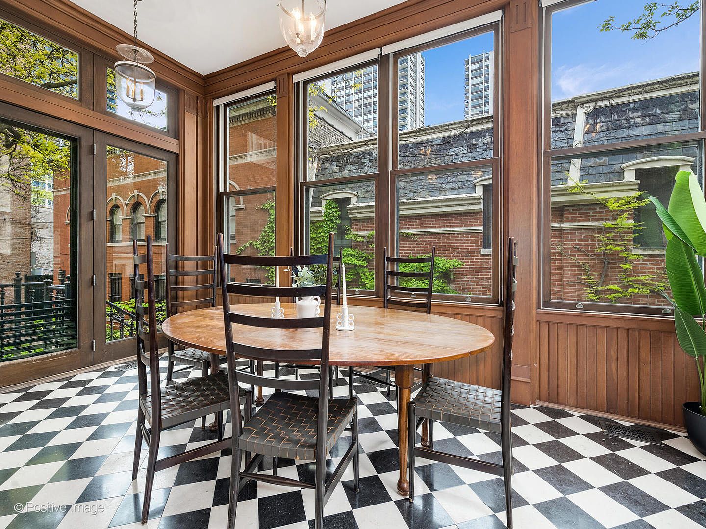 This sun-drenched dining area features a classic black-and-white checkered tile floor and rich wood-paneled walls that create a warm, traditional atmosphere. A round wooden table is centered in the room, surrounded by four dark-toned chairs, while large windows provide expansive views of the brick exterior and lush greenery outside. The space is illuminated by a hanging glass pendant light, offering a bright and inviting setting for meals.