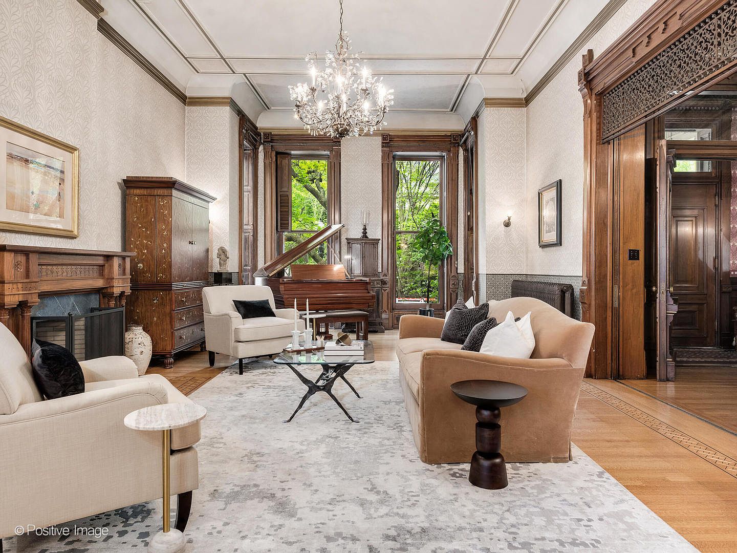 This elegant living room features a sophisticated blend of traditional architectural details, including ornate wood trim and a crystal chandelier, paired with contemporary furnishings. A plush beige sofa and cream-colored armchairs are arranged around a glass-topped coffee table on a large, patterned area rug. The space is anchored by a fireplace with a wood mantel and tall windows that provide a view of lush greenery outside.