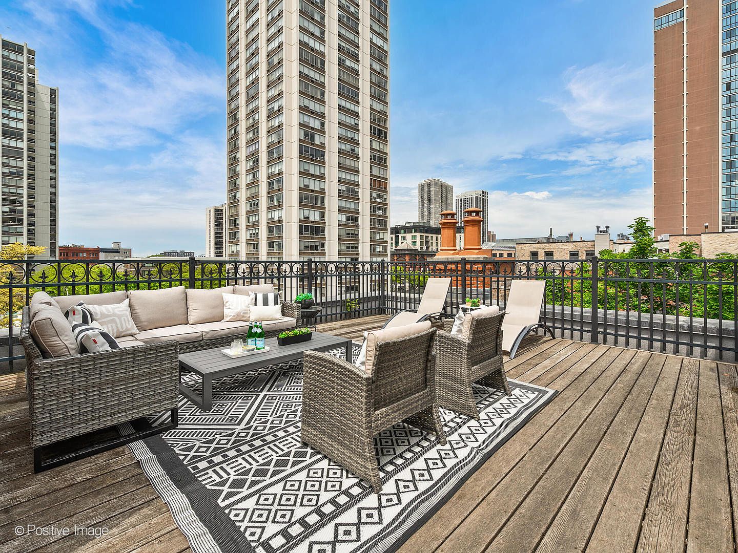 This expansive rooftop deck offers a sophisticated outdoor living space with a comfortable sectional sofa and matching armchairs arranged on a geometric patterned rug. The wooden decking provides a warm foundation, while a black metal railing ensures safety without obstructing the panoramic urban views of surrounding high-rise buildings. Two lounge chairs are positioned to take advantage of the open-air setting, creating an ideal environment for relaxation or entertaining in a city atmosphere.