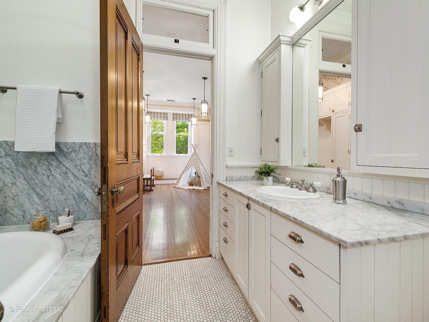 This bright bathroom features a white vanity with a stone countertop and a large mirror, complemented by a white tiled floor. A wooden door stands open, providing a clear view into an adjacent room that contains a play tent and hardwood flooring. The space feels clean and well-lit, with a classic aesthetic that transitions seamlessly into the living area.