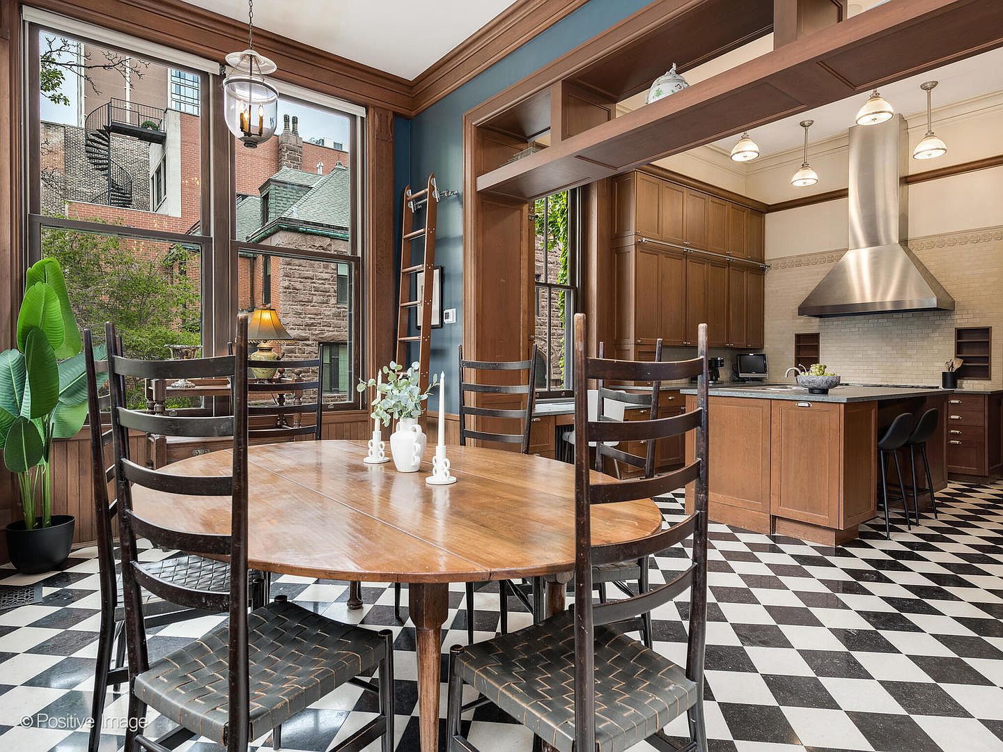This dining area features a round wooden table surrounded by high-backed chairs, set against a backdrop of large windows that offer views of neighboring brick buildings. The space is defined by a striking black-and-white checkered floor and deep blue wall accents, seamlessly transitioning into an open-concept kitchen with wood cabinetry and a stainless steel range hood. The perspective is eye-level, capturing the warm, traditional aesthetic of the dining space alongside the functional, modern design of the adjacent kitchen.