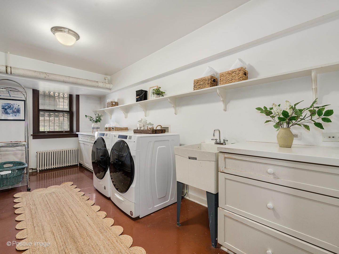 This bright and functional laundry room features a side-by-side washer and dryer set positioned next to a utility sink with a white cabinet base. The space includes open shelving for storage, a window with a view of the exterior, and a textured rug placed on the floor. The room is finished with white walls and a warm-toned floor, creating a clean and organized atmosphere.
