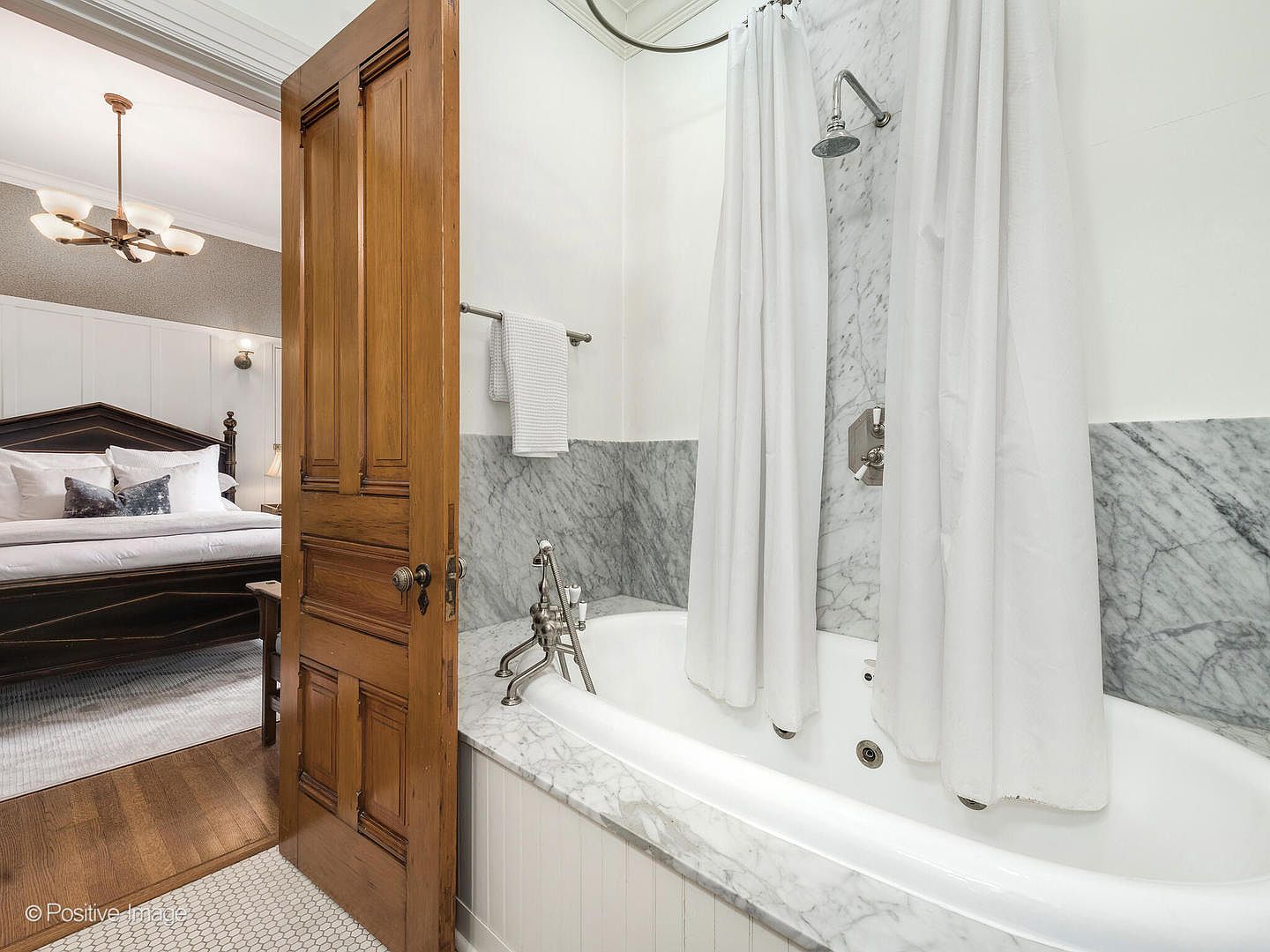 This bathroom features a jetted soaking tub integrated into a stone-topped deck with a shower head and white curtain. A classic wooden door stands slightly ajar, providing a view into an adjacent bedroom with a dark wood bed frame and a chandelier. The space combines traditional architectural elements with modern bathroom fixtures, creating a sophisticated and inviting atmosphere.