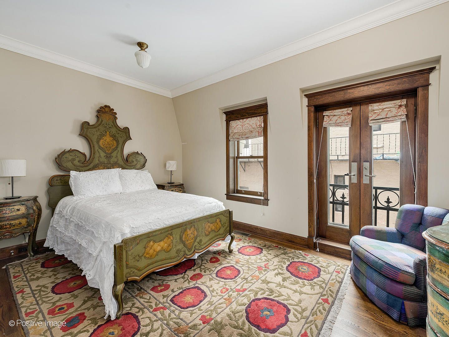 This bedroom features a distinctive, ornate painted wooden bed frame as the focal point, complemented by a large, colorful floral-patterned area rug. The room includes a cozy plaid armchair, dark wood-trimmed windows, and a set of French doors that open onto a small balcony. The neutral wall color provides a soft backdrop for the traditional, eclectic decor style.