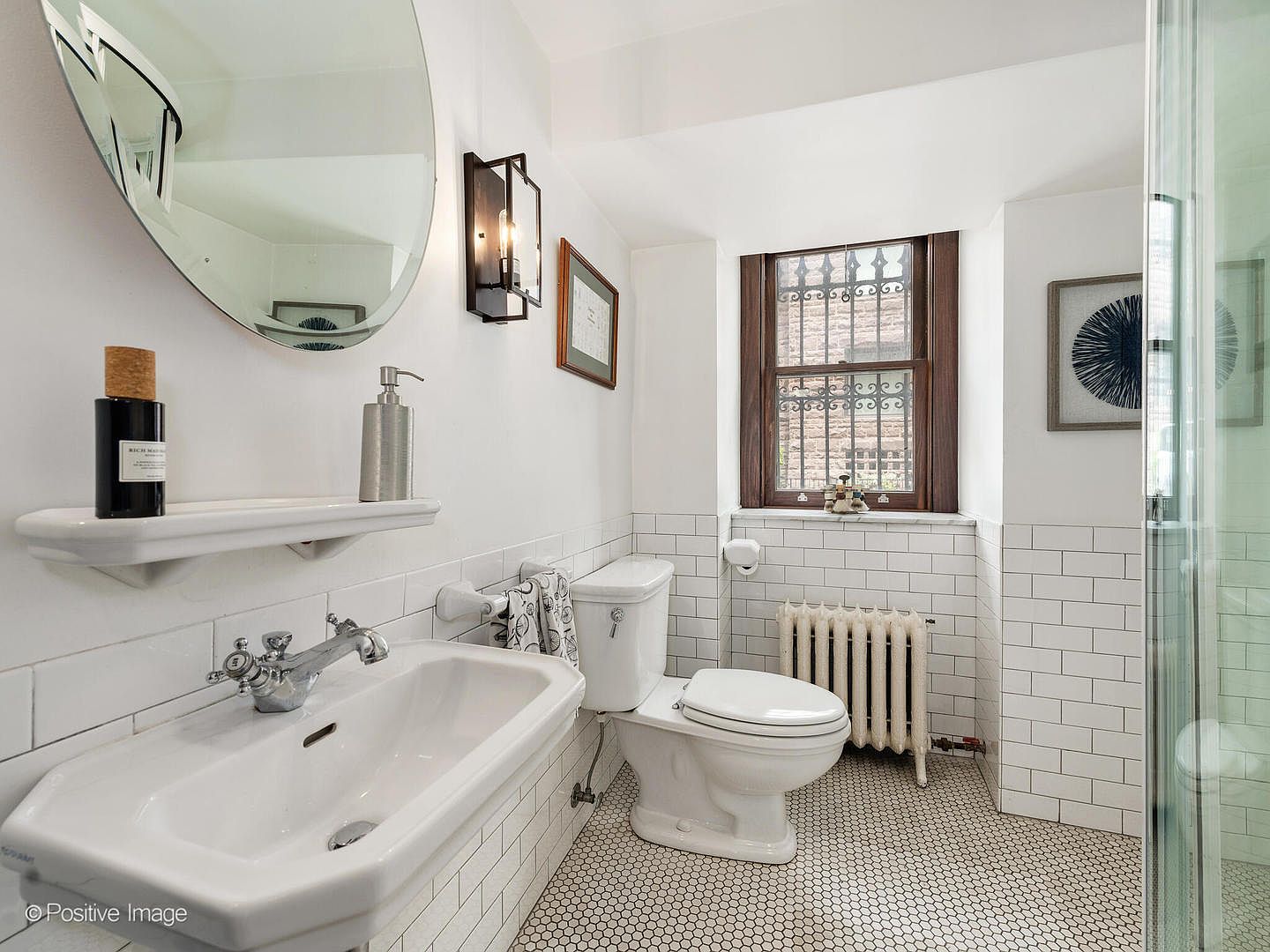 This bright, classic bathroom features white subway tile wainscoting and a distinctive black-and-white hexagonal tile floor. The space includes a pedestal sink with a chrome faucet, a white toilet, and a glass-enclosed shower, all illuminated by a wall-mounted lantern-style light fixture. A window with a dark wood frame provides natural light, while a round mirror above the sink adds a modern touch to the traditional aesthetic.