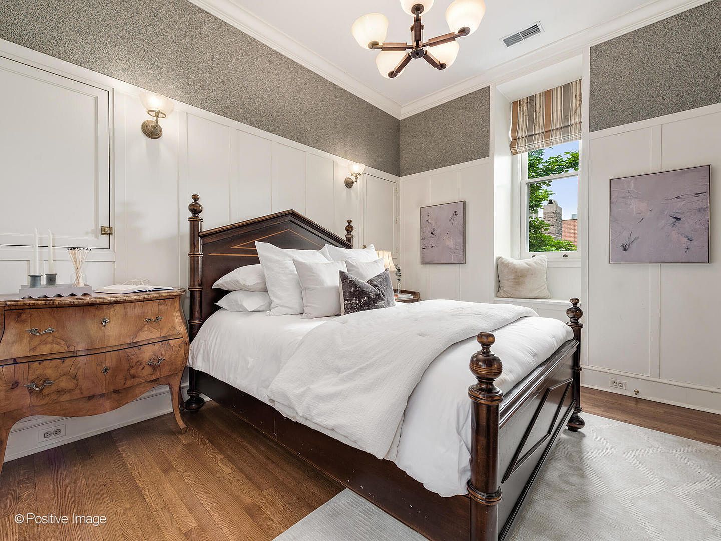 This elegant bedroom features a dark wood four-poster bed as the central focal point, dressed in crisp white linens and accented with decorative pillows. The room is characterized by white wainscoting on the lower walls and a textured, neutral-toned wallpaper on the upper portion, creating a sophisticated, classic aesthetic. A large window with a built-in bench seat provides natural light, while a vintage-style wooden dresser adds warmth and character to the space.