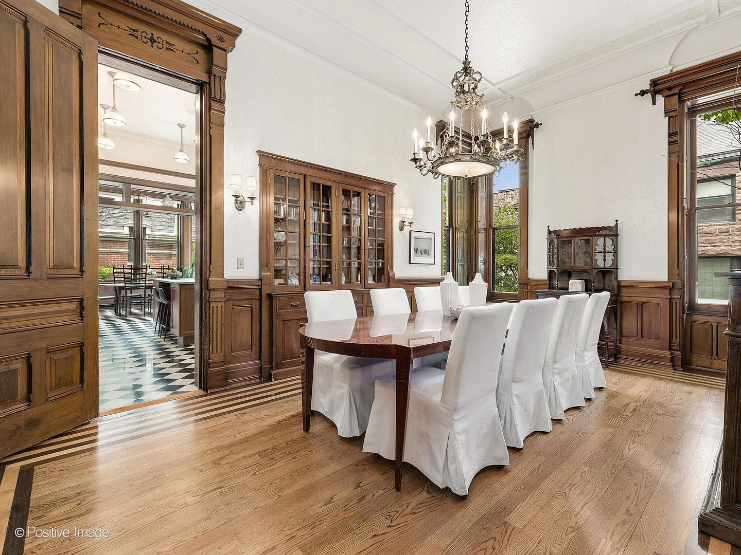 This elegant dining room features rich wood wainscoting and a large, oval dining table surrounded by eight white-slipcovered chairs. A grand, multi-armed chandelier hangs above the table, while a built-in wooden hutch provides ample storage and display space. The room opens into a bright, tiled kitchen area, showcasing a seamless flow between spaces with classic architectural details and warm wood flooring.