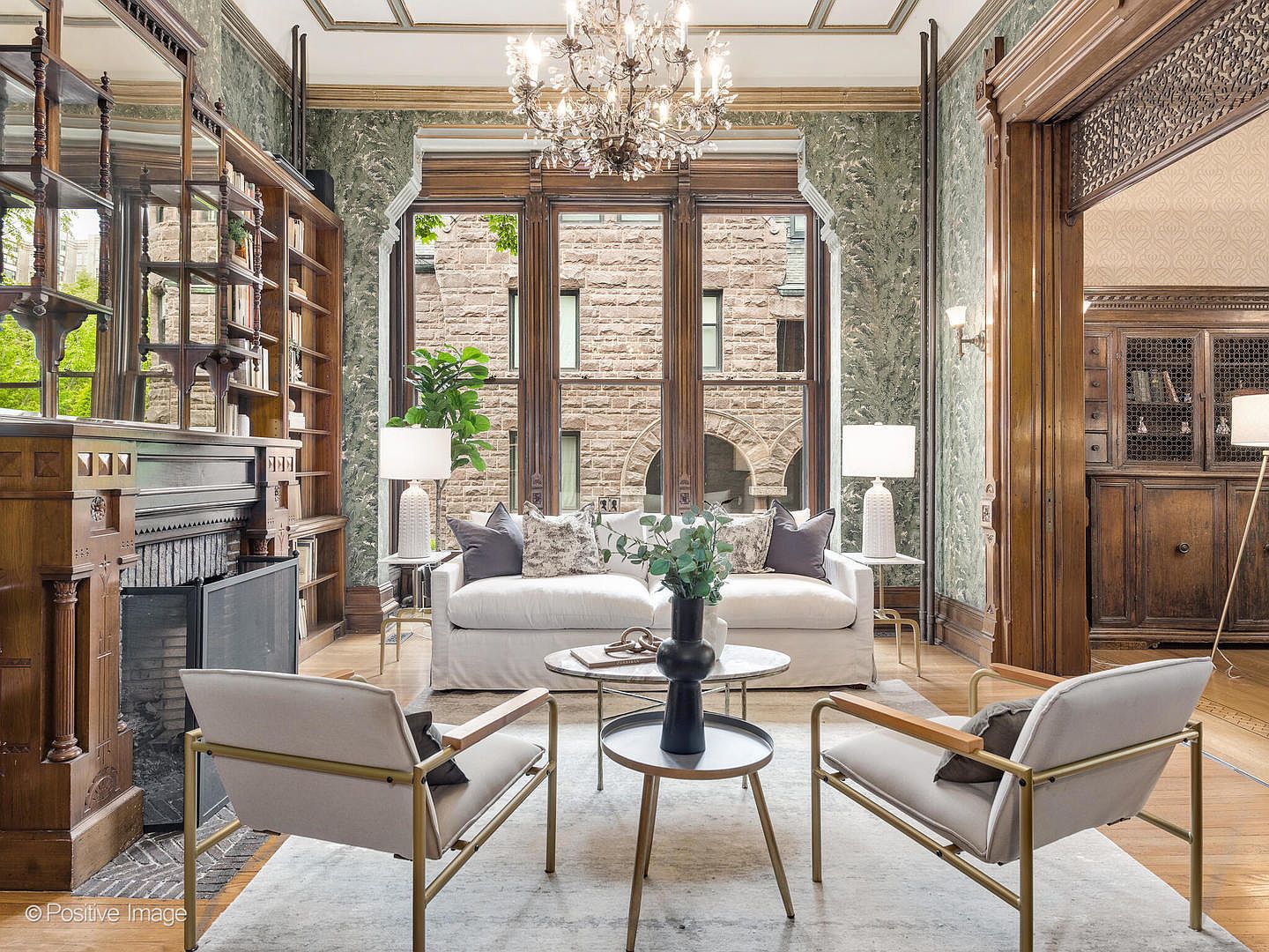 This elegant living room features a sophisticated blend of historic charm and modern comfort, highlighted by intricate wood paneling and a grand crystal chandelier. A plush white sofa is centered before a large window arrangement that frames a view of a stone building, while two contemporary armchairs and a tiered coffee table complete the seating area. The space is anchored by a fireplace with a detailed wood mantel and built-in shelving, creating a warm and inviting atmosphere.