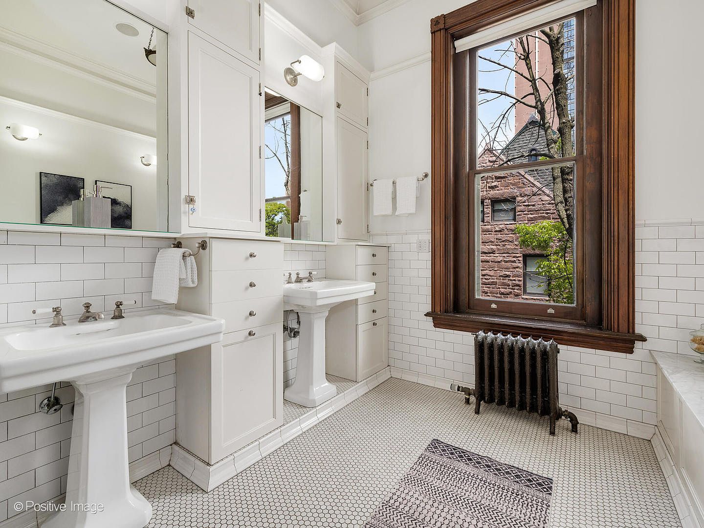 This bright, classic bathroom features dual pedestal sinks set against white subway-tiled walls and a mosaic-tiled floor. A large, dark-wood framed window provides natural light and a view of the exterior, while a vintage-style cast-iron radiator adds character to the space. The room is accented with built-in white cabinetry and mirrors, creating a clean, traditional aesthetic.