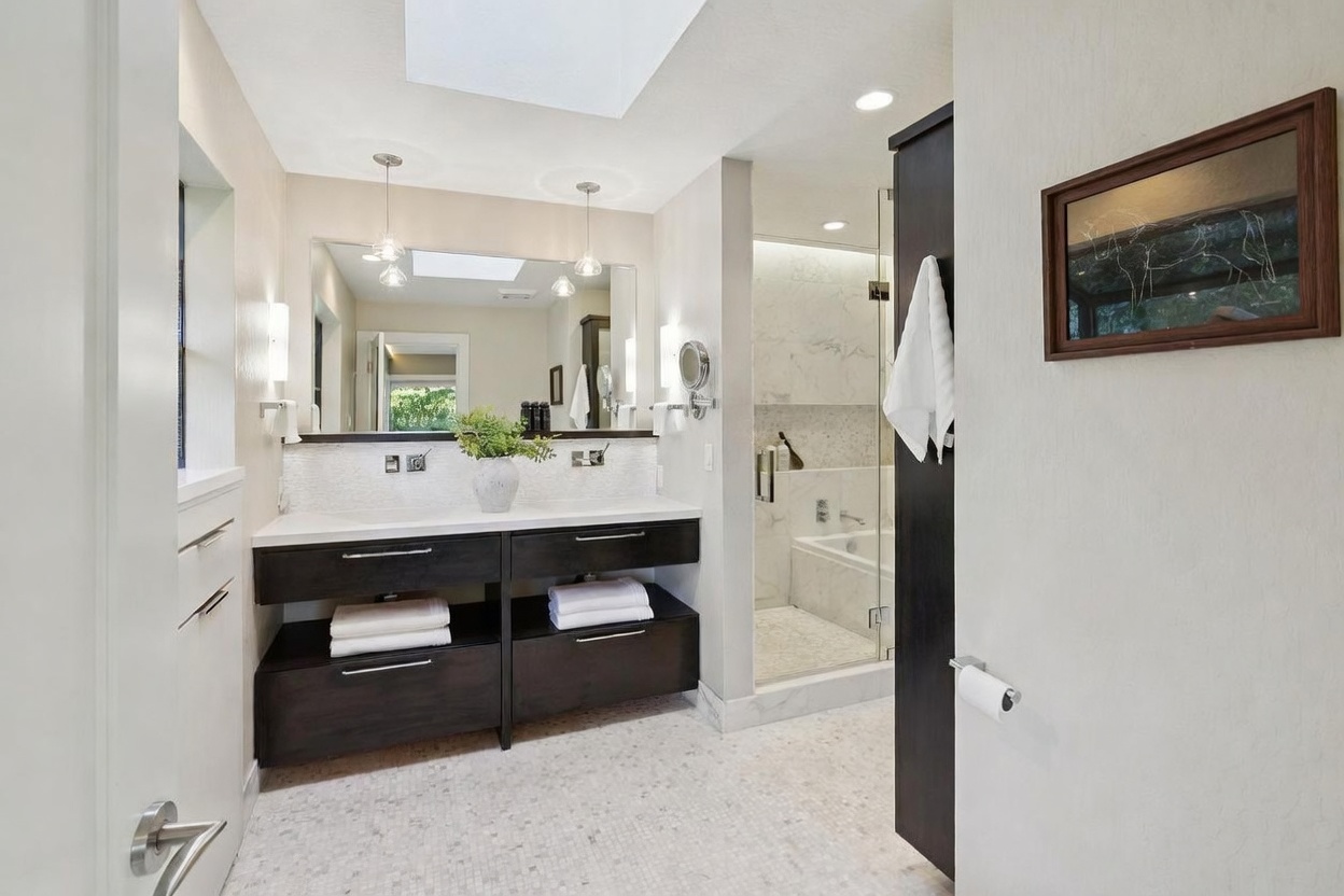 This modern primary bathroom features a sleek, dark wood double vanity with a white countertop and a large, frameless mirror illuminated by pendant lights. A glass-enclosed walk-in shower with marble tile work and a soaking tub is visible in the background, creating a spa-like atmosphere. The room is brightened by a skylight, and the overall aesthetic is clean, sophisticated, and contemporary.