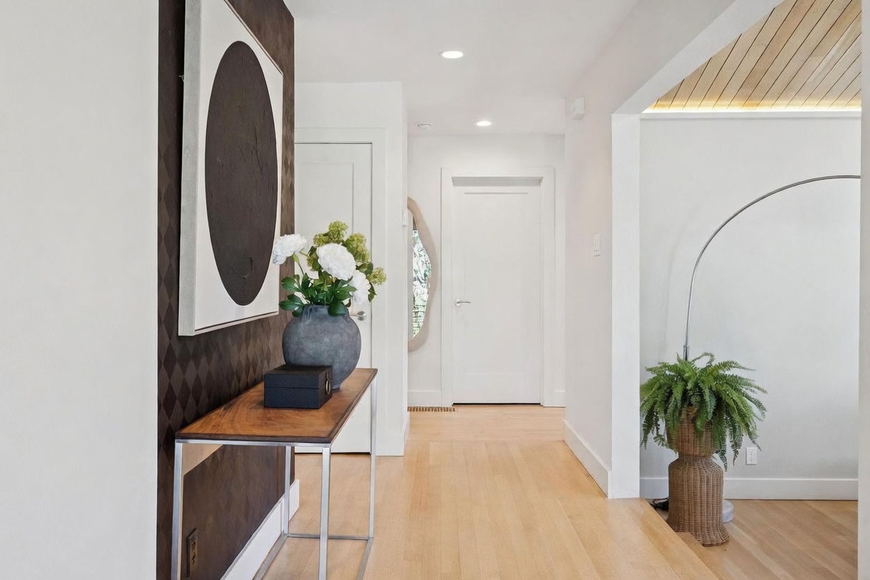 This bright and inviting entryway features light hardwood flooring and a modern aesthetic. A sleek console table sits against a textured, dark-patterned accent wall adorned with a large circular art piece, while a lush fern in a woven basket adds a touch of greenery to the space. The perspective is from the doorway looking into the home, highlighting the clean lines, recessed lighting, and a glimpse of a wood-paneled ceiling in the adjacent room.