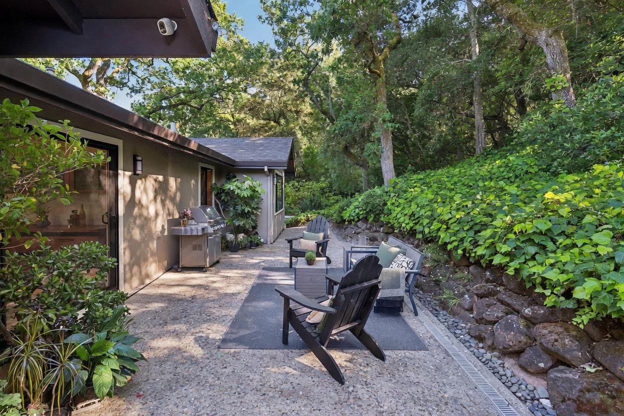This inviting outdoor patio space features a concrete surface furnished with comfortable Adirondack chairs and a small bench, perfect for relaxation. The area is bordered by a natural stone retaining wall covered in lush green ivy, creating a serene and private atmosphere. The perspective captures the transition from the home's exterior wall to the wooded backyard, highlighting a peaceful, shaded retreat ideal for outdoor dining or lounging.