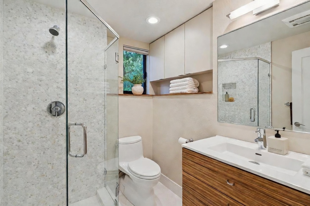 This modern bathroom features a sleek glass-enclosed shower with textured pebble-style tiling and a contemporary vanity with wood-grain cabinetry. A white toilet sits beside a window that brings in natural light, while built-in upper cabinets provide convenient storage above a wooden shelf. The overall aesthetic is clean, functional, and updated with a neutral color palette.