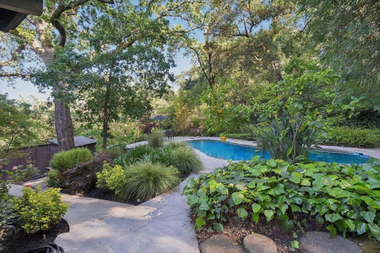 This serene backyard oasis features a sparkling blue swimming pool surrounded by lush, mature landscaping and towering oak trees. A stone pathway leads through the garden, while a small patio area with an umbrella provides a perfect spot for outdoor relaxation. The scene captures a peaceful, private retreat with a natural, wooded backdrop.