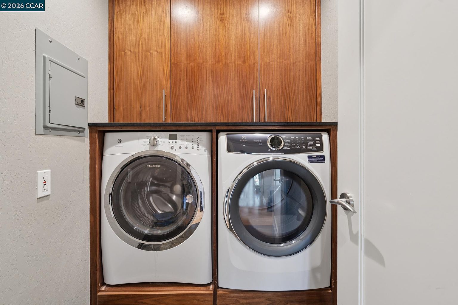 This laundry room features a sleek, built-in setup with a side-by-side Electrolux washer and dryer set nestled under a dark countertop. Above the appliances, warm-toned wood cabinetry provides ample storage, creating a clean and organized aesthetic. The space is efficiently designed, incorporating a wall-mounted electrical panel and a neutral color palette that feels both functional and modern.