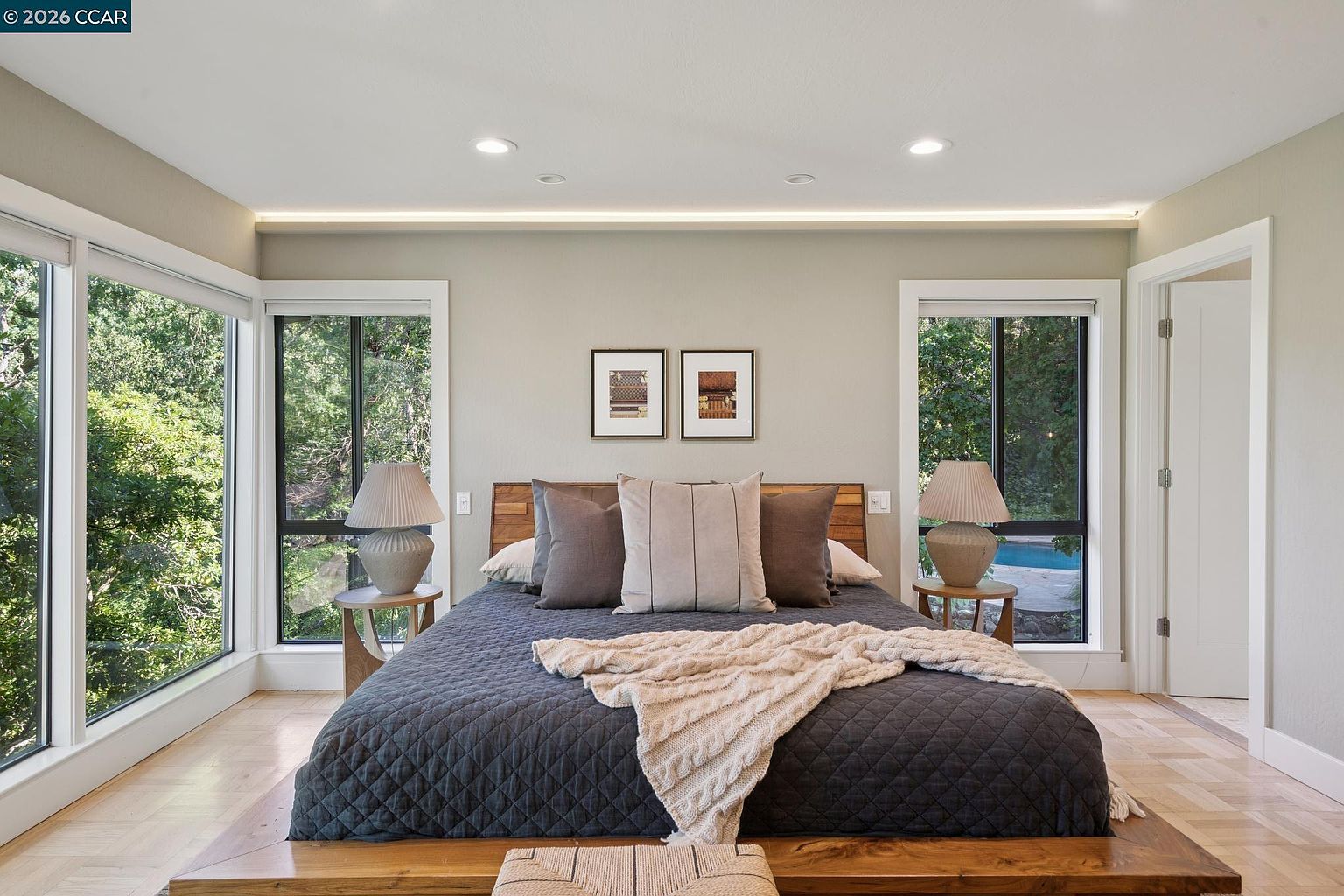 This primary bedroom features a modern, minimalist aesthetic with a low-profile wooden platform bed frame and a dark quilted comforter. Large windows on either side of the bed provide abundant natural light and views of the surrounding greenery and a glimpse of a pool, while a soft, neutral color palette creates a serene atmosphere. The room is accented by symmetrical bedside tables with textured lamps and two framed art pieces centered above the headboard.