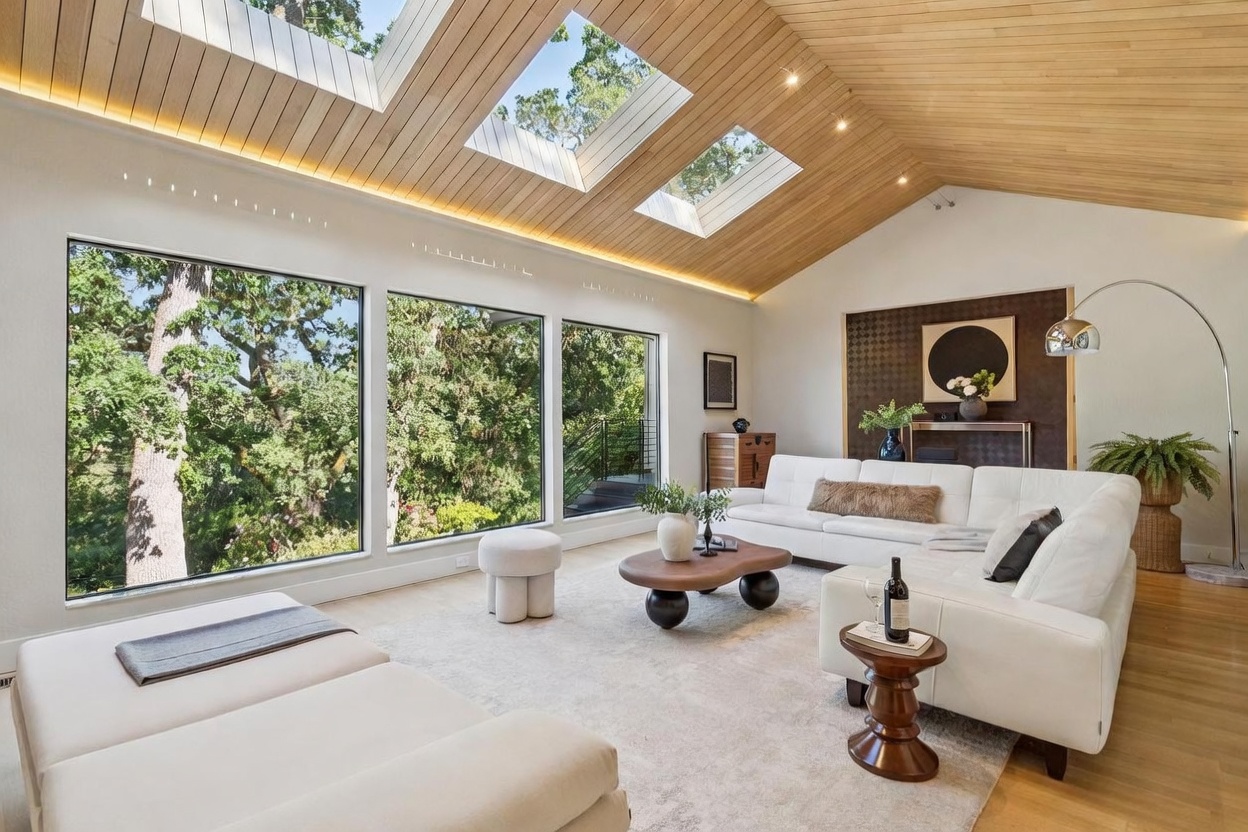 This bright and airy living room features a dramatic vaulted wood-paneled ceiling with large skylights that flood the space with natural light. A plush white sectional sofa anchors the room, complemented by a unique organic-shaped coffee table and expansive windows that frame a lush, tree-filled outdoor view. The space exudes a sophisticated, modern aesthetic with warm wood tones and a neutral color palette, creating an inviting and serene atmosphere.