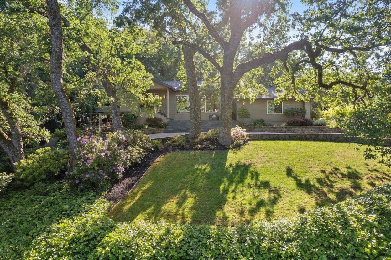 This image captures a charming, multi-level home nestled among mature oak trees, showcasing a blend of natural beauty and architectural appeal. The front yard features a lush, manicured lawn, vibrant purple rhododendrons, and a stone retaining wall that leads up to the house entrance. The perspective is a wide, eye-level shot that emphasizes the home's integration into its wooded, serene environment.