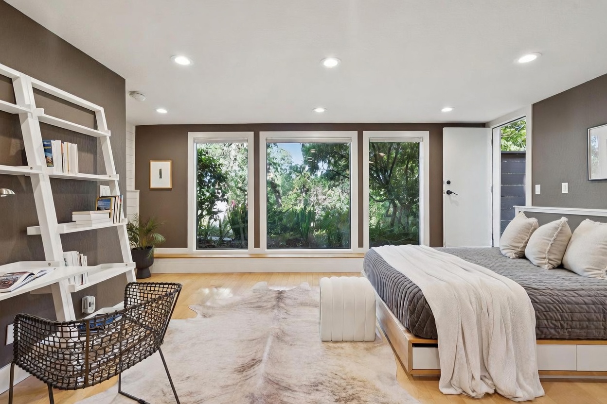 This bright and airy primary bedroom features a modern aesthetic with dark accent walls, light hardwood flooring, and a large, plush bed dressed in neutral tones. A unique ladder-style shelving unit adds a contemporary touch, while a wall of floor-to-ceiling windows provides a serene view of the lush greenery outside. The room is anchored by a textured area rug and a minimalist white ottoman, creating a calm and inviting retreat.