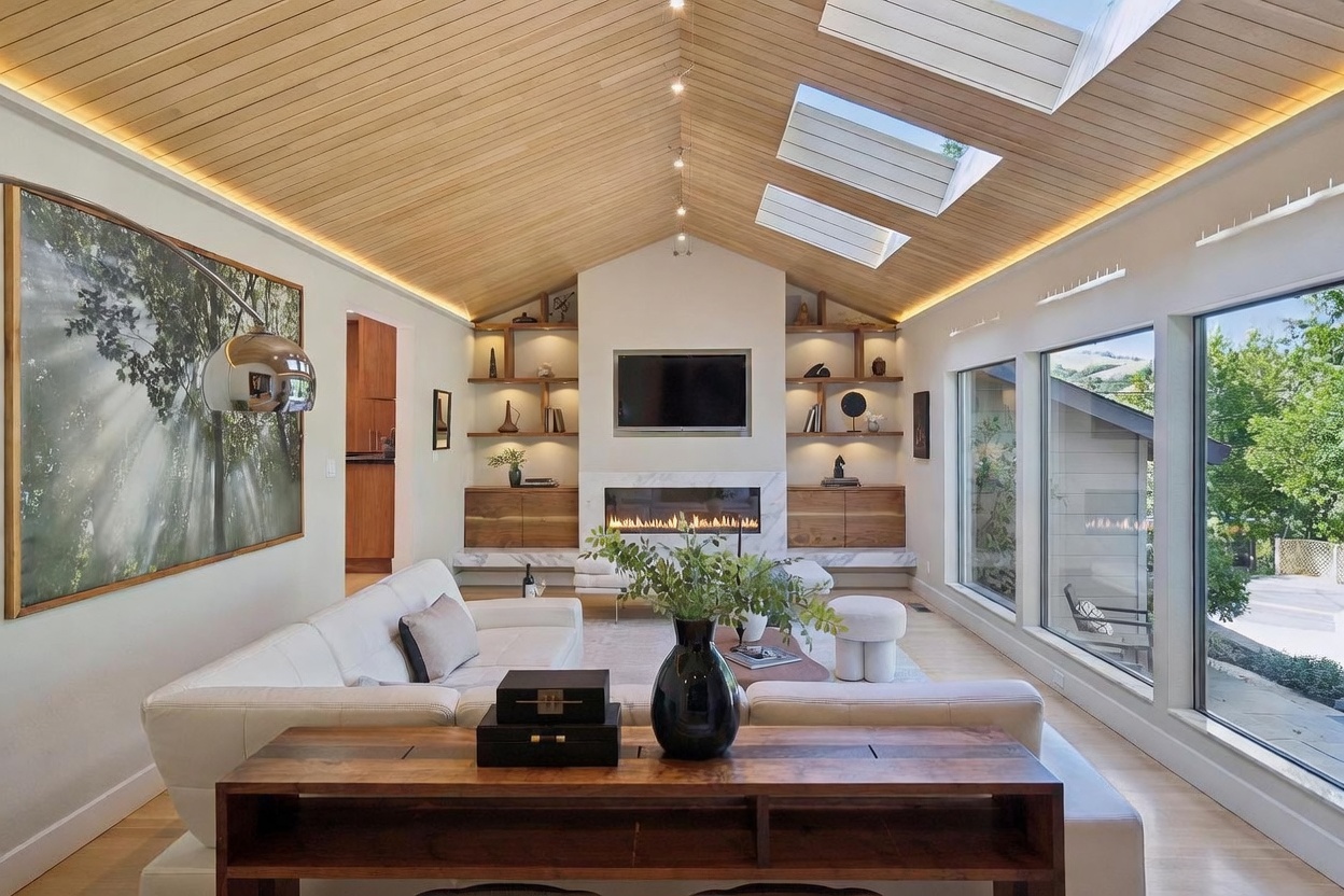 This sophisticated living room features a dramatic vaulted ceiling with warm wood paneling and integrated recessed lighting. The space is anchored by a sleek, linear fireplace set into a marble surround, flanked by custom built-in shelving and cabinetry. Large floor-to-ceiling windows and skylights flood the room with natural light, creating an airy, modern atmosphere that seamlessly blends indoor comfort with outdoor views.