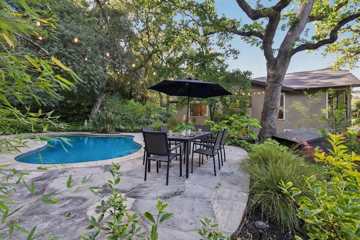 This inviting backyard oasis features a kidney-shaped swimming pool set against a backdrop of lush, mature trees and greenery. A stone patio provides a perfect outdoor dining area, complete with a table, chairs, and a black umbrella, creating a serene and private atmosphere. The scene is captured from a ground-level perspective, highlighting the seamless blend of natural landscaping and functional outdoor living space.