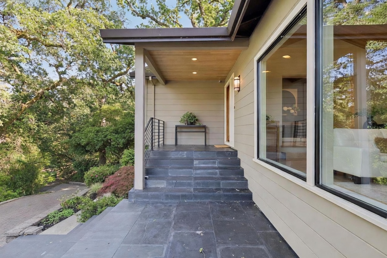 This inviting entryway features a set of dark slate stairs leading up to a covered porch with a warm wood-paneled ceiling and recessed lighting. A sleek black metal railing lines the steps, while a minimalist console table with a potted plant sits near the front door. The exterior is finished in neutral horizontal siding, and a large window provides a glimpse into the bright, modern interior of the home.