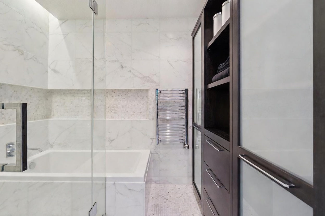 This luxurious primary bathroom features a sleek, modern design with floor-to-ceiling marble tiling and a deep soaking tub. A chrome heated towel rack adds a functional touch of elegance, while a tall, dark wood storage unit with glass-front cabinets provides sophisticated organization. The perspective captures the clean lines and high-end finishes, creating an atmosphere of a spa-like retreat.
