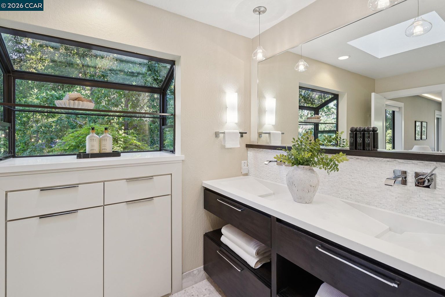 This modern primary bathroom features a sleek, dark wood vanity with a white integrated double sink countertop and a textured tiled backsplash. A large, frameless mirror reflects the space, while a wide window above the adjacent storage cabinet provides natural light and a view of the surrounding greenery. The room is accented with contemporary light fixtures and clean, minimalist lines, creating a sophisticated and serene atmosphere.