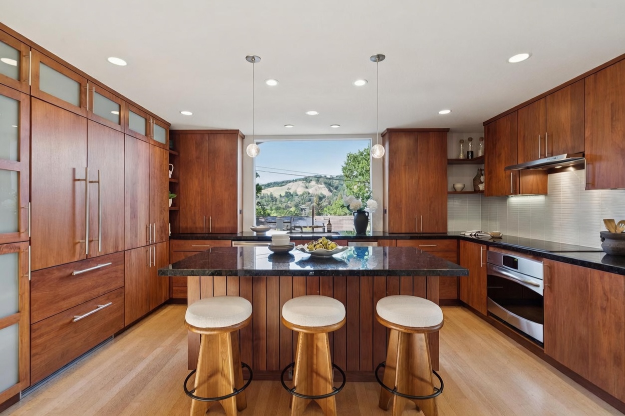This modern kitchen features warm-toned wood cabinetry, sleek black granite countertops, and a central island with three bar stools. A large window above the sink provides a scenic view of the hills, while pendant lights add a touch of elegance to the space. The overall aesthetic is sophisticated and inviting, blending natural wood textures with contemporary finishes.