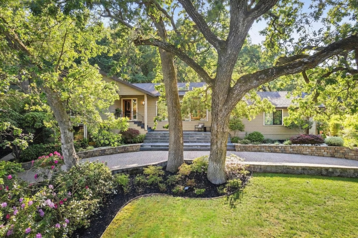 This inviting single-story home features a warm, earthy-toned exterior nestled among mature oak trees and lush landscaping. A wide, stone-paved circular driveway leads to a grand set of steps ascending to the front entrance, creating a stately and welcoming curb appeal. The scene is captured from a ground-level perspective, emphasizing the harmonious blend of natural foliage and the home's architectural design.