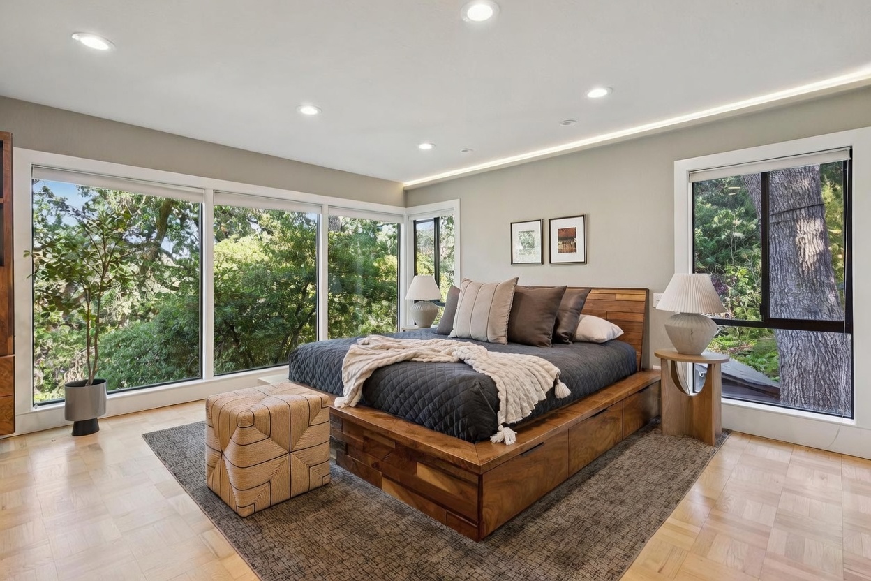 This serene primary bedroom features a modern wooden platform bed with built-in storage, centered against a wall with large windows that provide an immersive view of the surrounding trees. The room is decorated in a neutral, earthy palette with a textured area rug, a geometric ottoman, and minimalist bedside lamps, creating a tranquil and sophisticated atmosphere. The perspective is a wide, eye-level shot that emphasizes the seamless connection between the interior space and the natural landscape outside.