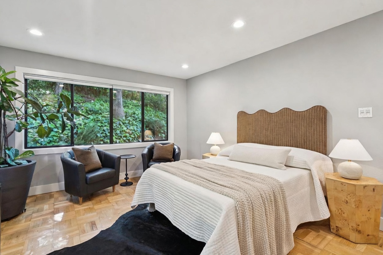 This bright and airy primary bedroom features a textured, scalloped-edge headboard and a plush white bed dressed with a cozy knit throw. The room is accented by two sleek black leather armchairs positioned near a large window that offers a serene view of the lush, wooded backyard. The space is completed with light wood parquet flooring, a dark area rug, and warm bedside lighting, creating a sophisticated and tranquil retreat.