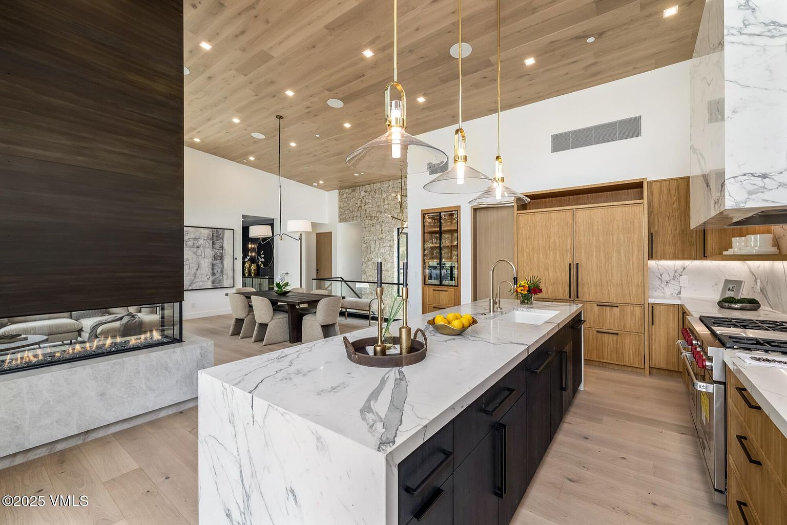 This open-concept kitchen features a large central island with a stone countertop and dark cabinetry, complemented by light wood cabinetry along the perimeter. A tall, dark-paneled fireplace surround anchors the space, while a dining area with a wooden table and upholstered chairs is visible in the background. The room is illuminated by three glass pendant lights and recessed lighting, set against a light wood-paneled ceiling that adds warmth to the modern design.