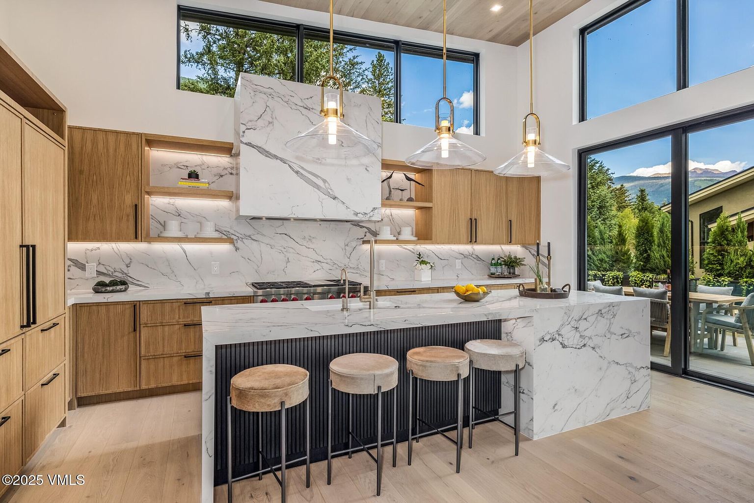This modern kitchen features a large central island with a stone countertop and a dark, slatted base, complemented by four upholstered bar stools. The space is defined by light wood cabinetry, open shelving, and a prominent stone-clad range hood that anchors the cooking area. Large windows above the cabinets and a sliding glass door provide abundant natural light and views of the surrounding landscape, creating an airy and sophisticated atmosphere.