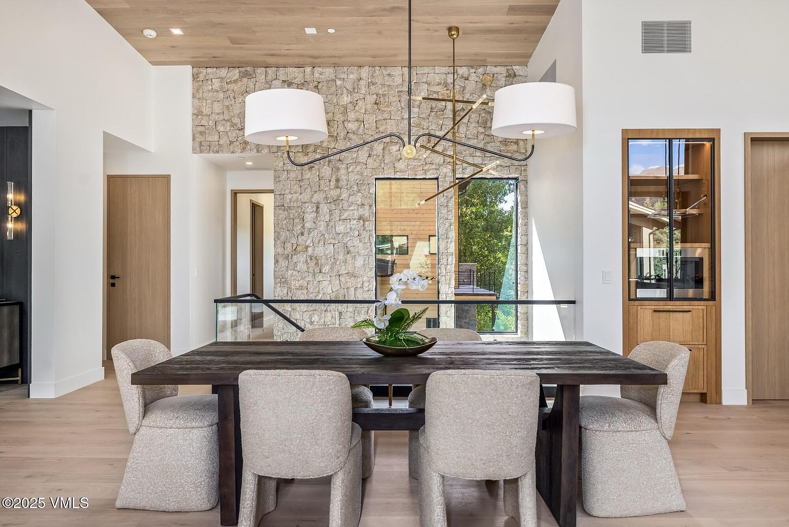 This elegant dining room features a dark, rustic wood table surrounded by four upholstered chairs in a neutral, textured fabric. A striking, modern multi-arm chandelier hangs above the table, centered against a dramatic wall clad in light-toned stacked stone. The space is framed by light wood ceilings and a glass-paneled railing that overlooks a lower level, creating an open and sophisticated atmosphere.
