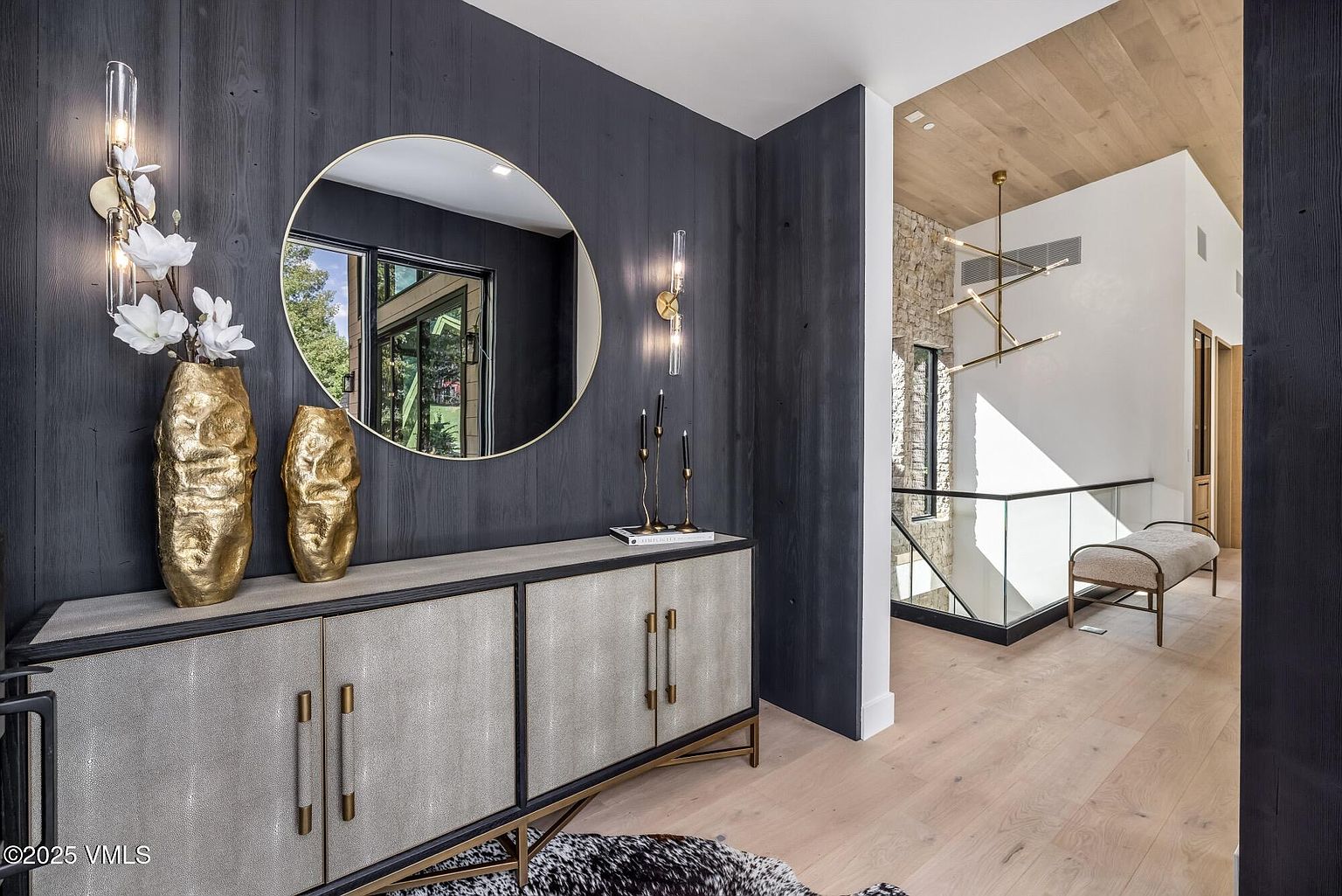 This sophisticated entryway features a striking dark wood-paneled wall accented by a large circular mirror and gold-toned wall sconces. A textured console table sits below, complemented by gold decorative vases, while the adjacent space reveals a staircase with a glass railing and a view of a stone-clad wall. The light wood flooring and modern chandelier create a warm, high-end atmosphere that flows into the open-concept home.