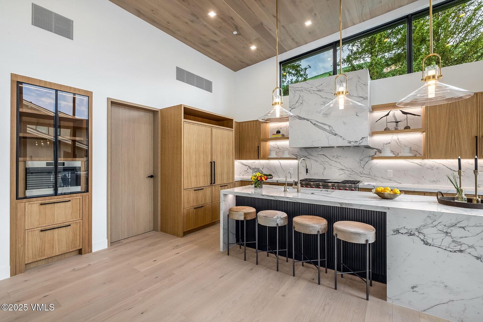 This modern kitchen features a large central island with a stone countertop and a dark, slatted base, complemented by four upholstered bar stools. The space is defined by light wood cabinetry, a striking stone-clad range hood, and open shelving illuminated by integrated lighting. Large windows above the cooking area allow natural light to flood the room, which is finished with a light wood ceiling and matching flooring.