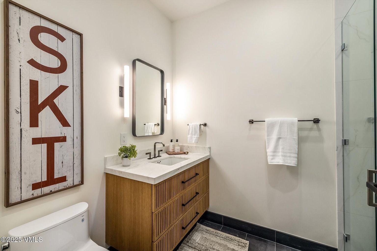 This modern bathroom features a floating vanity with a slatted wood finish and a light-colored stone countertop. A rectangular mirror with integrated side lighting hangs above the sink, while a large, rustic-style sign reading 'SKI' adds a decorative touch to the wall. The space is completed by dark floor tiles and a glass-enclosed shower, creating a clean and contemporary aesthetic.