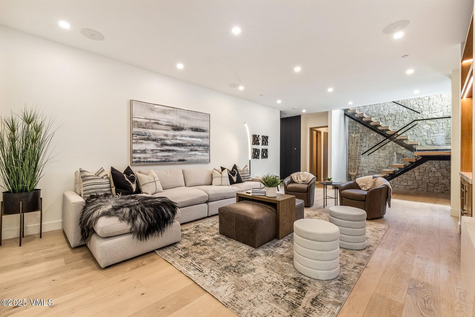 This contemporary living room features a large, light-colored sectional sofa adorned with decorative pillows and a faux-fur throw, centered around a leather ottoman and a wooden coffee table. The space is anchored by a textured area rug and complemented by two leather armchairs and two round, fabric-covered ottomans. A striking staircase with a stone-clad wall serves as a dramatic backdrop, while recessed lighting and a large abstract painting enhance the sophisticated, open-concept design.