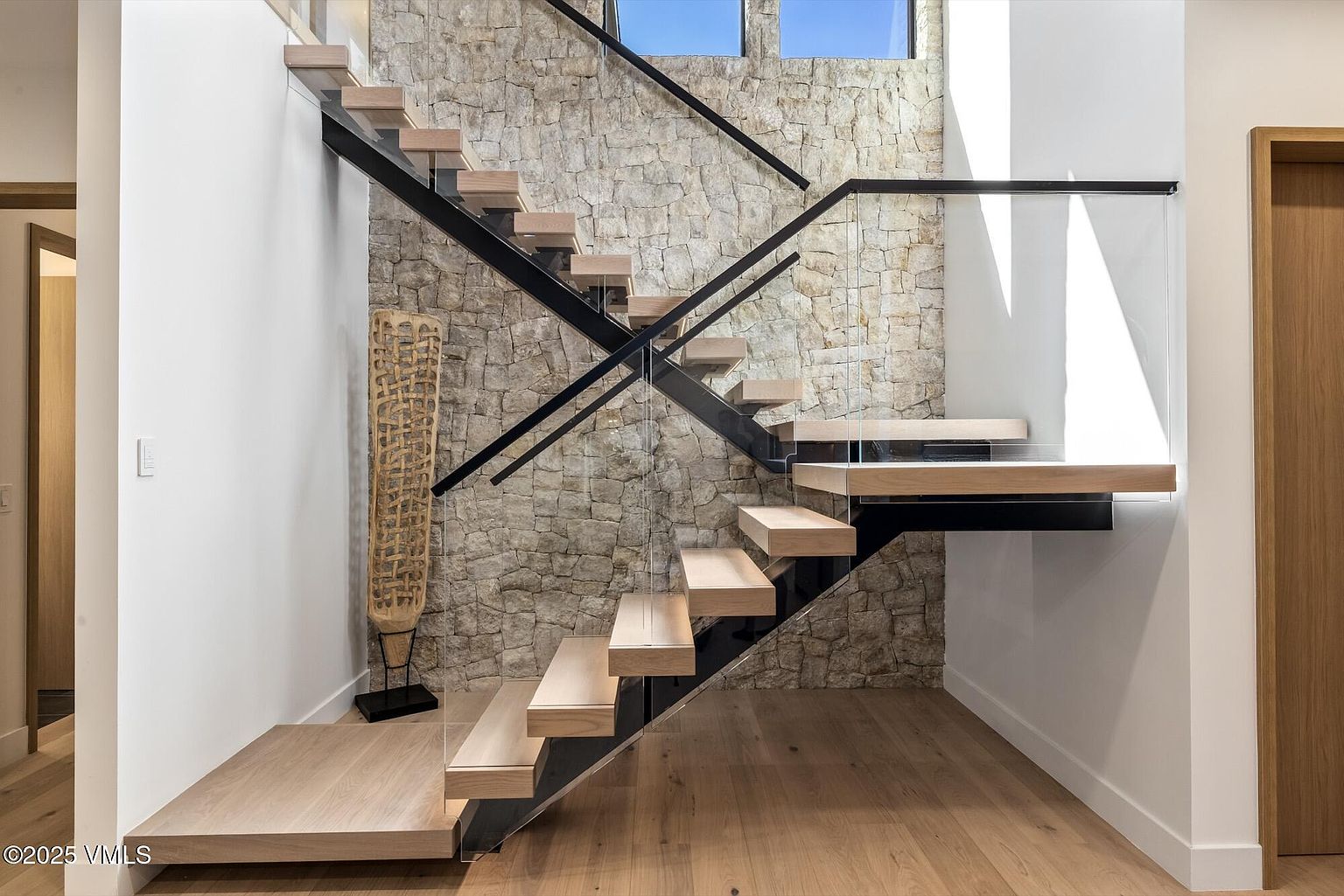 This striking interior space features a modern floating staircase with light wood treads and a sleek black metal frame. The stairs are set against a textured stone wall that adds depth and organic character to the contemporary design, while a glass railing maintains an open and airy feel. A tall, decorative woven sculpture stands at the base of the stairs, enhancing the sophisticated and architectural aesthetic of the entryway.