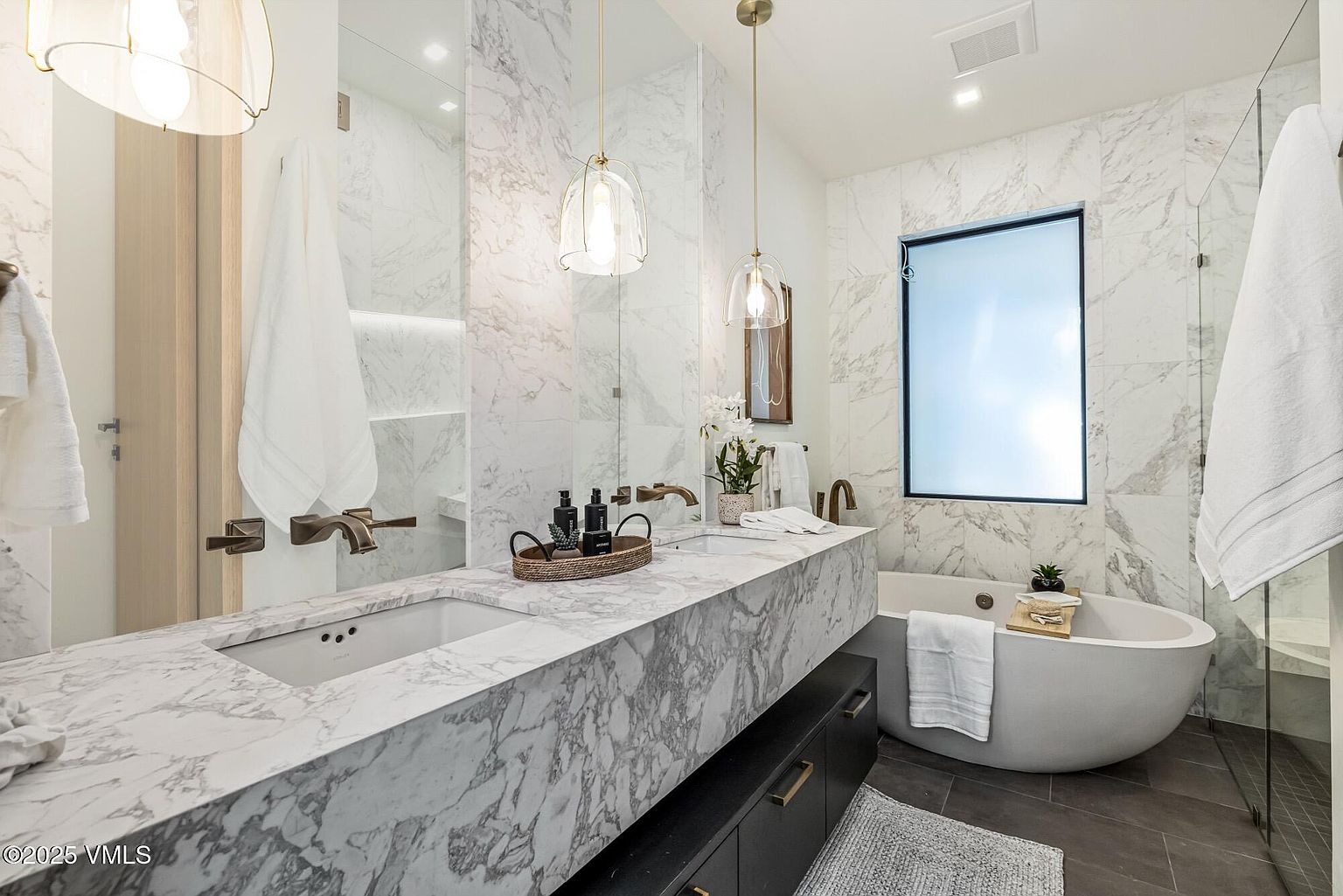 This luxurious primary bathroom features a floating vanity with a striking stone countertop and dual wall-mounted faucets. A freestanding soaking tub sits beneath a large frosted window, complemented by a glass-enclosed shower area and elegant pendant lighting. The space is finished with large-format stone wall tiles and dark cabinetry, creating a sophisticated and modern aesthetic.