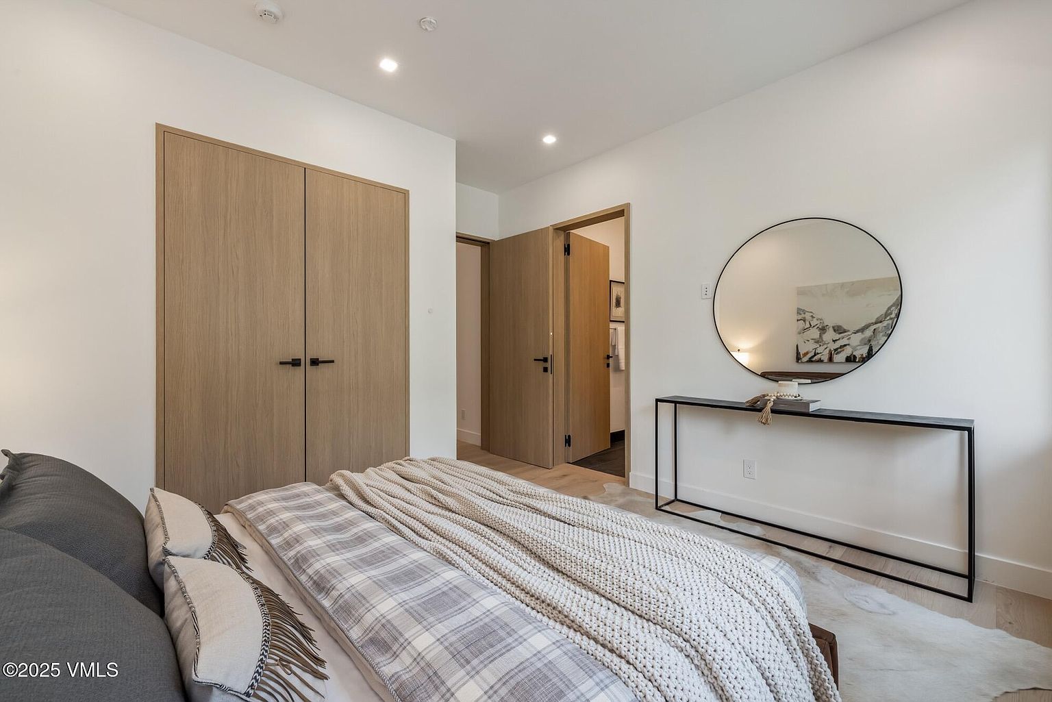 This contemporary bedroom features a neutral color palette with light wood-toned doors and flooring. A bed dressed in a plaid duvet and a chunky knit throw serves as the focal point, while a minimalist black console table with a large circular mirror adds a modern touch to the wall. The perspective is from the foot of the bed, looking toward the closet and an open doorway leading to an adjacent room.