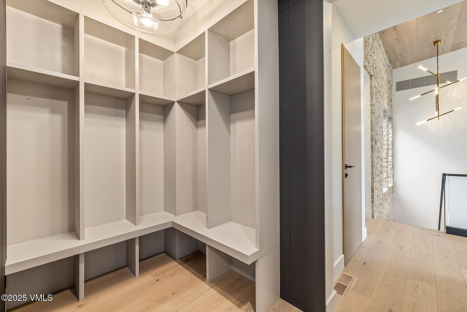 This image showcases a custom-built walk-in closet or mudroom area featuring light-toned, built-in shelving and bench seating arranged in an L-shape. The space is illuminated by a modern, multi-bulb light fixture, and the light wood flooring creates a cohesive, airy aesthetic. To the right, a doorway leads into an adjacent space with a stone-clad wall and a contemporary chandelier, suggesting a sophisticated and well-designed home interior.