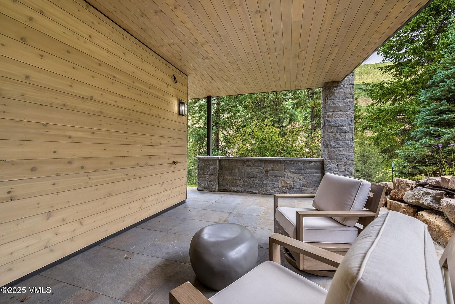 This covered patio features a modern aesthetic with light wood paneling on the walls and ceiling, complemented by stone accents and large stone floor tiles. Two comfortable armchairs with light-colored cushions face a low stone wall that overlooks a lush, wooded landscape. The space offers a serene, private outdoor retreat with a seamless blend of natural materials and contemporary design.
